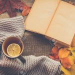 Tasse Tee, gemütlicher gestrickter Schal, Herbstlaub, offenes Buch und  Kürbis auf Holzbrett.